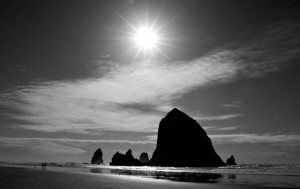 Black and white photograph of rock protrusions on the Pacific coast; couple waling on the beach; sun setting.