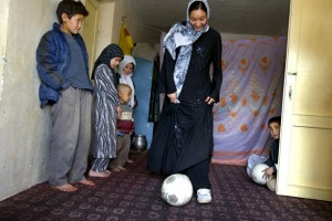 Young woman wearing a hajib and dribbling a soccer ball while her siblings look on.